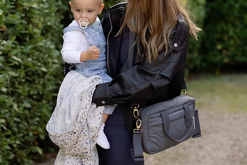 Woman holding a baby and carrying a gray handbag outdoors.