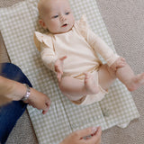 Baby in a pink outfit lying on a checkered blanket with a person's hands nearby.