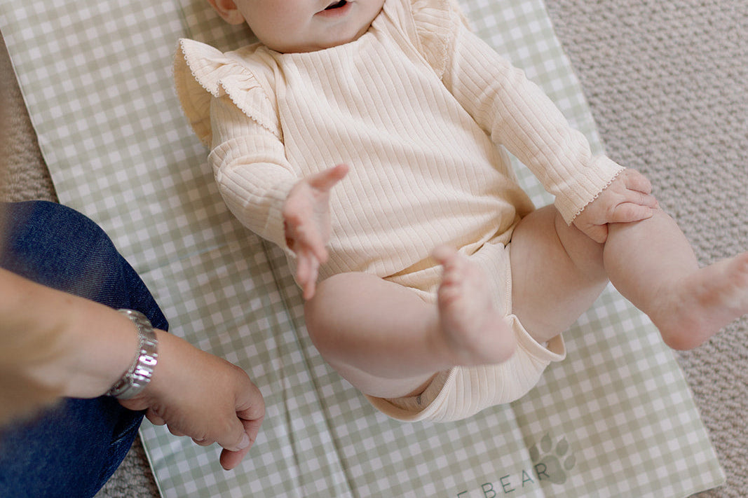 Baby in a pink outfit lying on a checkered blanket with a person's hands nearby.