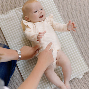 Baby in a white onesie lying on a checkered blanket with an adult's hands nearby.