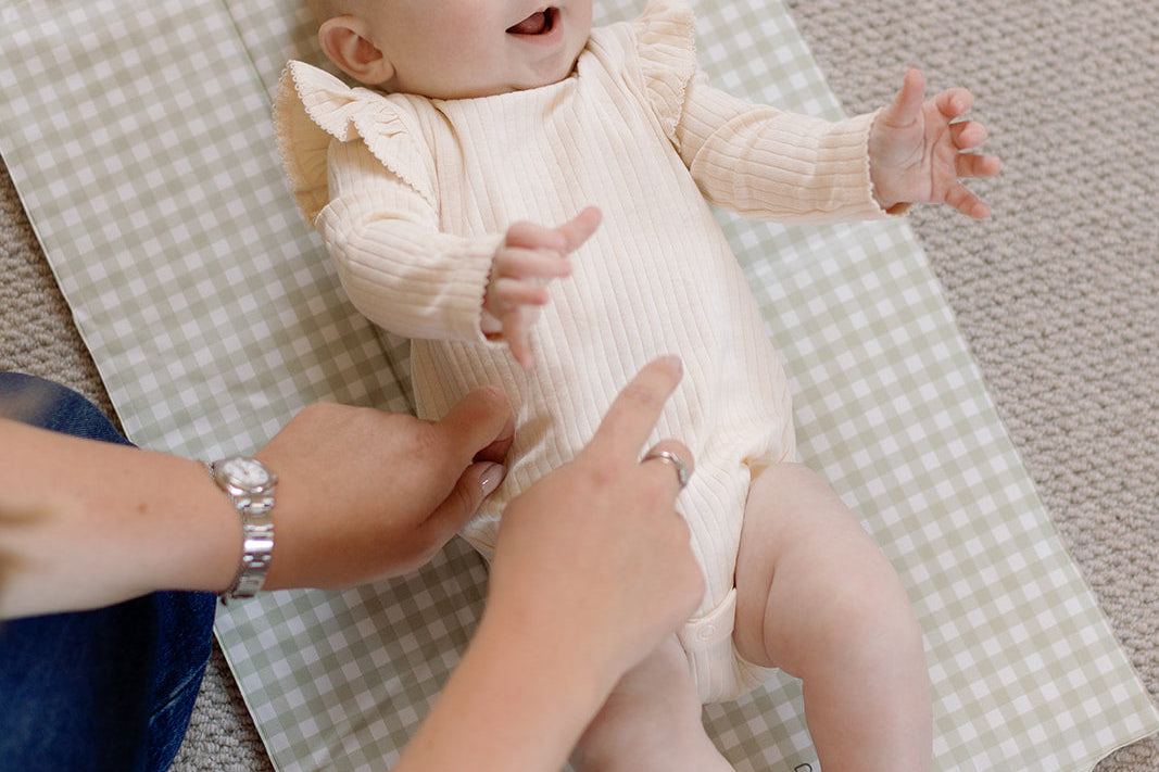 Baby in a white onesie lying on a checkered blanket with an adult's hands nearby.