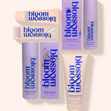 A set of seven Bloom & Assoi skincare products arranged on a light cream background. The products include tubes, bottles, and boxes in lavender purple and pale peach-beige colors with bold blue branding.