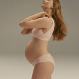 Pregnant model wearing a light pink maternity bra and matching underwear, posing in profile against a neutral gray studio background.