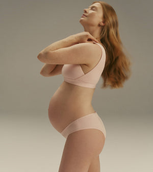Pregnant model wearing a light pink maternity bra and matching underwear, posing in profile against a neutral gray studio background.