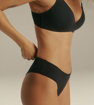 A close-up side view of a model wearing a solid black two-piece bikini set against a neutral light gray studio background.