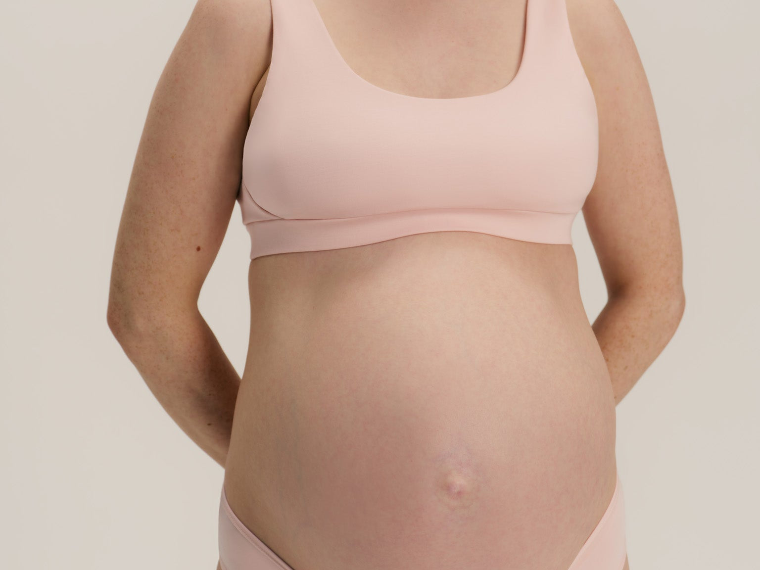 A pregnant woman wearing a light pink maternity bra and matching underwear, standing against a neutral light gray background.