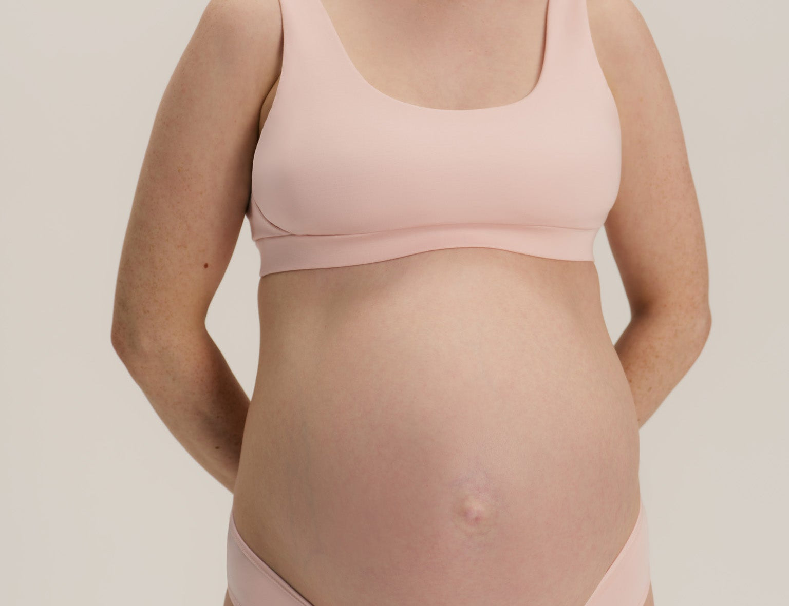 A pregnant woman wearing a light pink maternity bra and matching underwear, standing against a neutral light gray background.