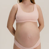 A pregnant woman wearing a light pink maternity bra and matching underwear, standing against a neutral light gray background.