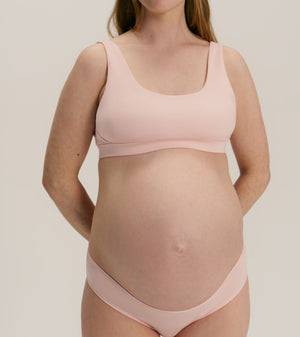 A pregnant woman wearing a light pink maternity bra and matching underwear, standing against a neutral light gray background.
