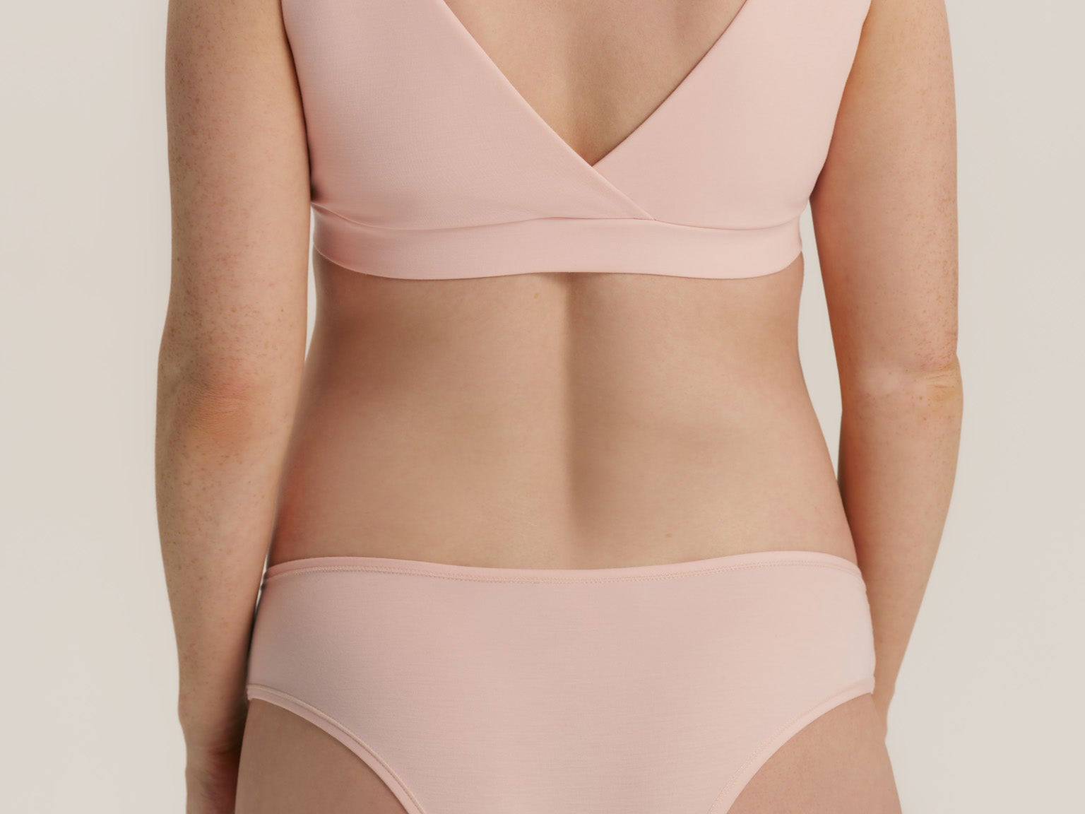 A model shown from the back wearing a light pink two-piece bikini set, featuring a wrap-style bra top and matching high-cut briefs, against a solid light beige background.