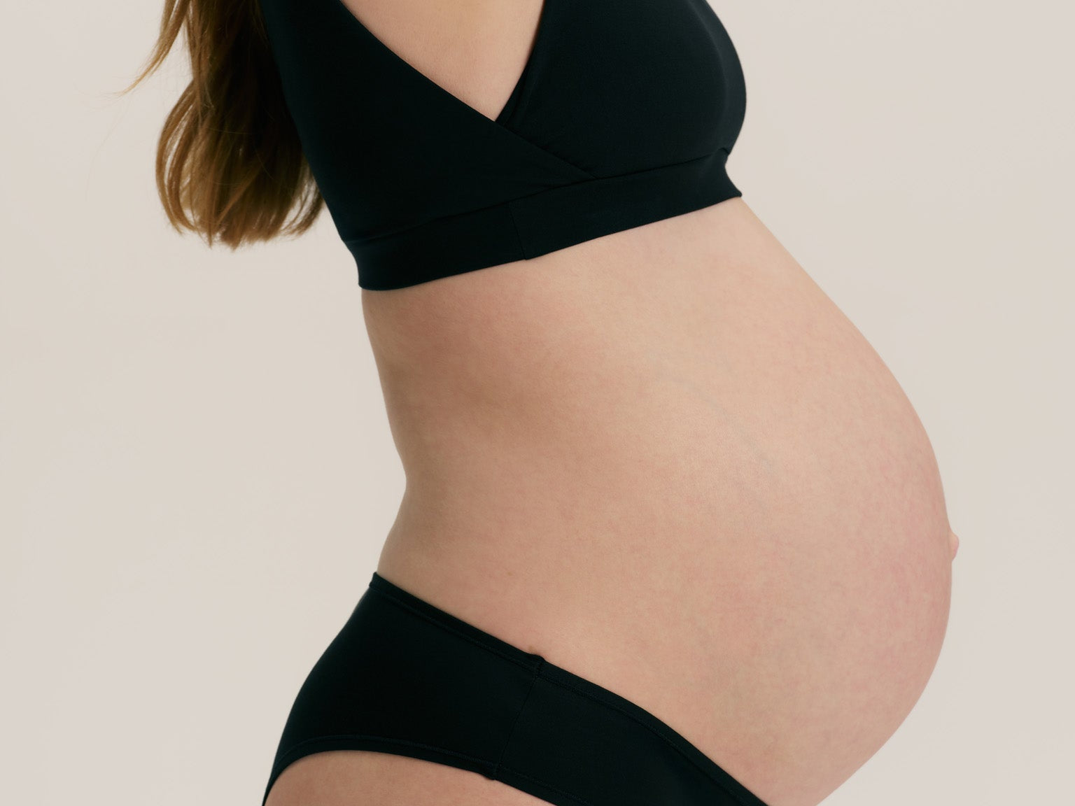 Pregnant model wearing a black maternity bralette and matching high-waisted underwear bottoms against a solid light beige background.
