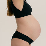 Pregnant model wearing a black maternity bralette and matching high-waisted underwear bottoms against a solid light beige background.