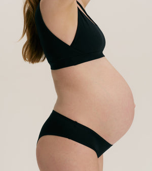 Pregnant model wearing a black maternity bralette and matching high-waisted underwear bottoms against a solid light beige background.