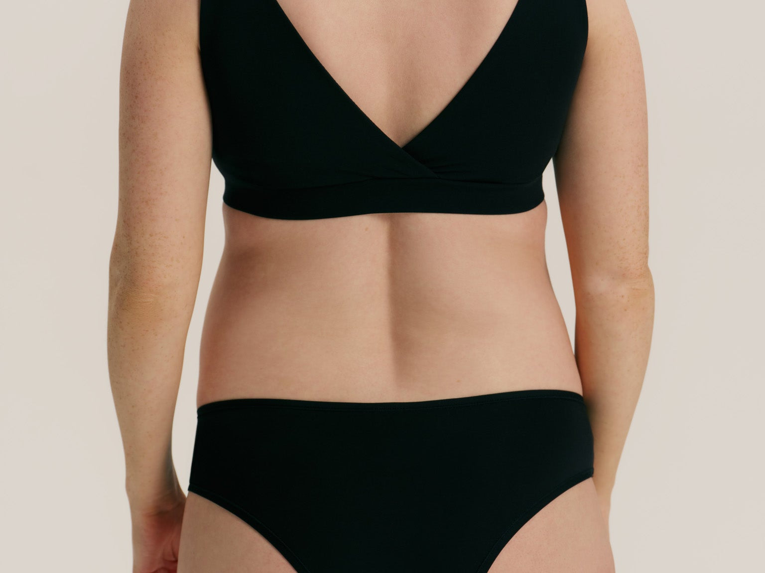 Rear view of a model wearing a black two-piece bikini set with a wrap-style bra top and high-cut bottoms against a plain light beige studio background.