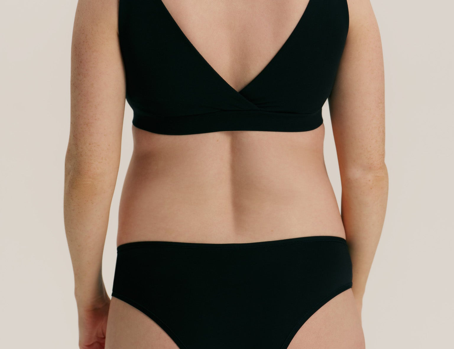 Rear view of a model wearing a black two-piece bikini set with a wrap-style bra top and high-cut bottoms against a plain light beige studio background.