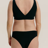 Rear view of a model wearing a black two-piece bikini set with a wrap-style bra top and high-cut bottoms against a plain light beige studio background.