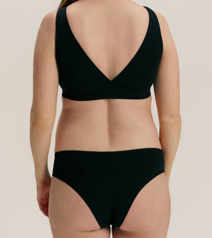 Rear view of a model wearing a black two-piece bikini set with a wrap-style bra top and high-cut bottoms against a plain light beige studio background.