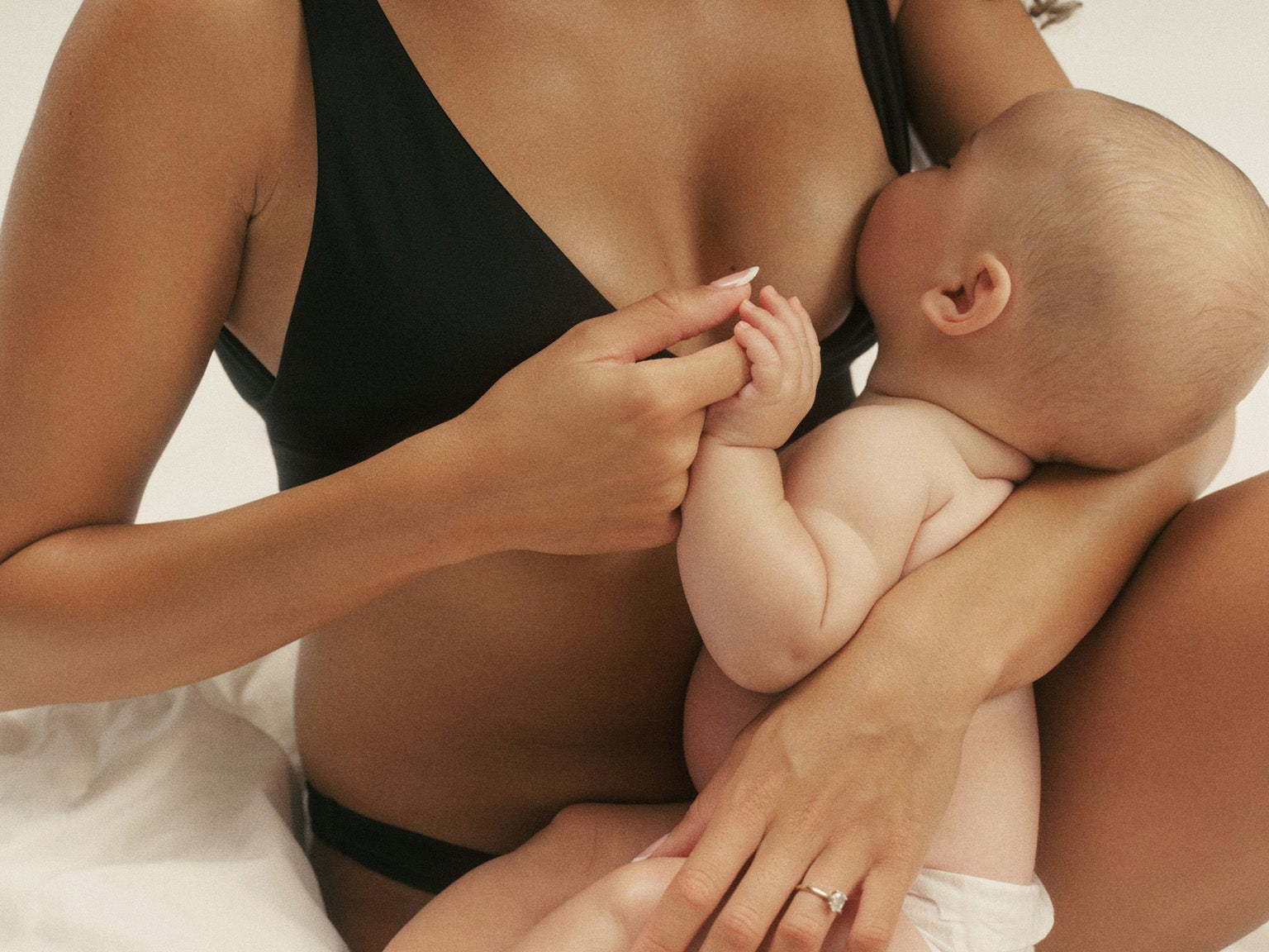 A woman in a black nursing bra breastfeeding a baby while sitting on white bedding. The woman has curly dark hair and is wearing a black bra, and the baby is wearing a white diaper.