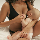 A woman in a black nursing bra breastfeeding a baby while sitting on white bedding. The woman has curly dark hair and is wearing a black bra, and the baby is wearing a white diaper.
