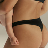 Close-up of a person wearing black underwear, with a baby held against the torso. The shot is cropped tightly and focuses on the underwear in a studio setting with a light neutral background.