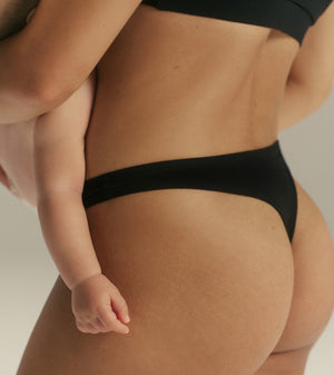 Close-up of a person wearing black underwear, with a baby held against the torso. The shot is cropped tightly and focuses on the underwear in a studio setting with a light neutral background.