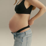 Pregnant woman wearing a black maternity support bra and light blue jeans, shown from the side against a neutral studio background.