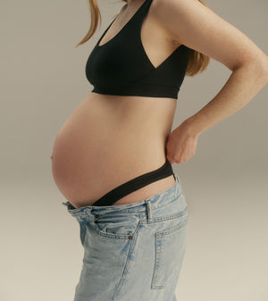 Pregnant woman wearing a black maternity support bra and light blue jeans, shown from the side against a neutral studio background.