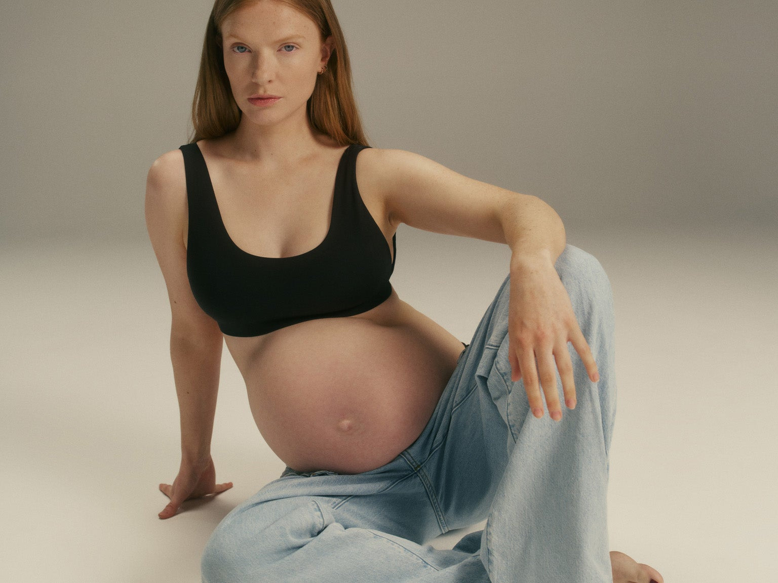 A pregnant woman with red hair sits on the floor in a studio setting, wearing a black tank top and light blue jeans.