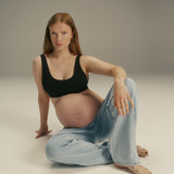 A pregnant woman with red hair sits on the floor in a studio setting, wearing a black tank top and light blue jeans.