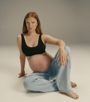A pregnant woman with red hair sits on the floor in a studio setting, wearing a black tank top and light blue jeans.
