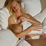 A blonde woman in white nursing lingerie sits on a bed with white pillows and bedding, breastfeeding a baby wearing a white bodysuit. The brand name 'nuna' is visible on the baby's bodysuit.