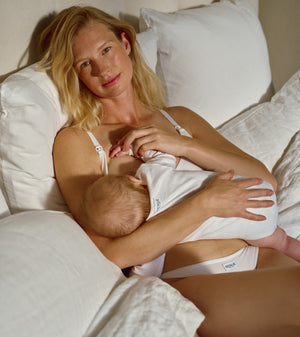 A blonde woman in white nursing lingerie sits on a bed with white pillows and bedding, breastfeeding a baby wearing a white bodysuit. The brand name 'nuna' is visible on the baby's bodysuit.