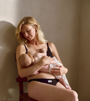 A woman in black underwear sits on a wooden chair, breastfeeding a baby wrapped in a white blanket in a softly lit indoor setting.