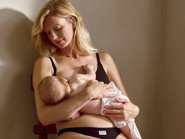 A woman in black underwear sits on a wooden chair, breastfeeding a baby wrapped in a white blanket in a softly lit indoor setting.