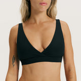 A close-up of a woman wearing a black wrap-style underwire bra against a plain light background.