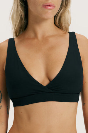 A close-up of a woman wearing a black wrap-style underwire bra against a plain light background.