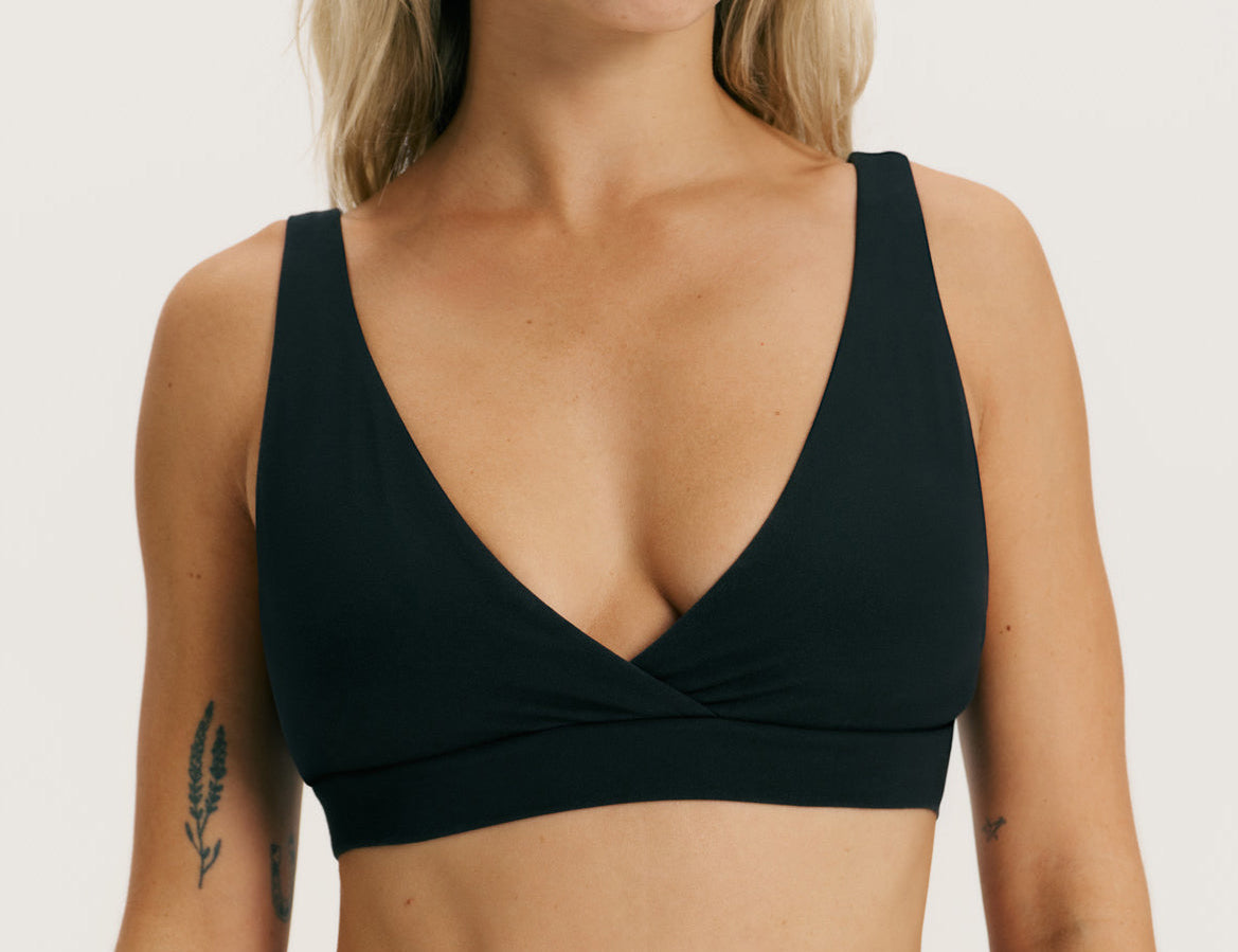 A close-up of a woman wearing a black wrap-style underwire bra against a plain light background.