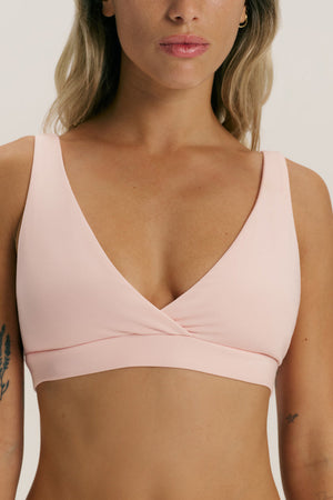 A woman wearing a light pink wrap-style sports bra against a plain light gray studio background.