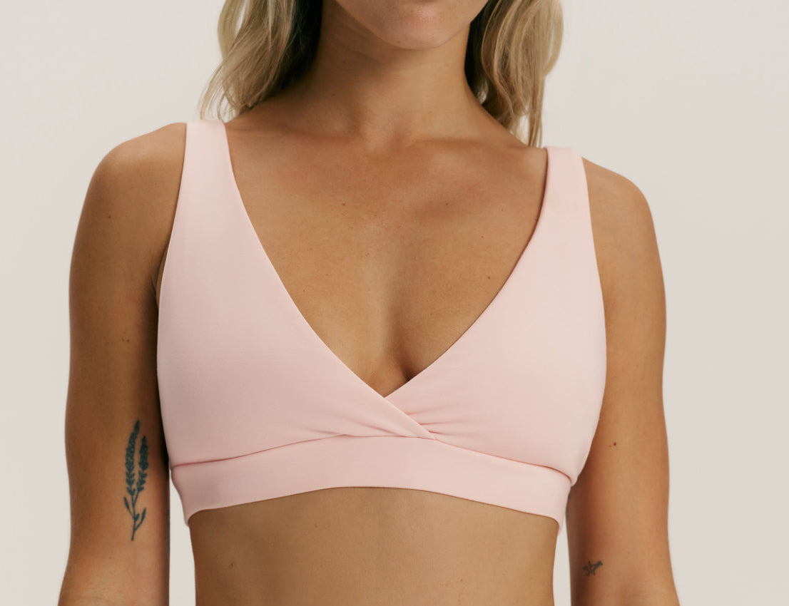 A woman wearing a light pink wrap-style sports bra against a plain light gray studio background.