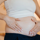 A close-up lifestyle shot of a pregnant woman wearing a white ribbed tank top and dark navy pants, cradling her baby bump with her hands. She is wearing gold rings, and the background is a soft, warm beige.