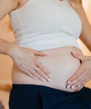 A close-up lifestyle shot of a pregnant woman wearing a white ribbed tank top and dark navy pants, cradling her baby bump with her hands. She is wearing gold rings, and the background is a soft, warm beige.
