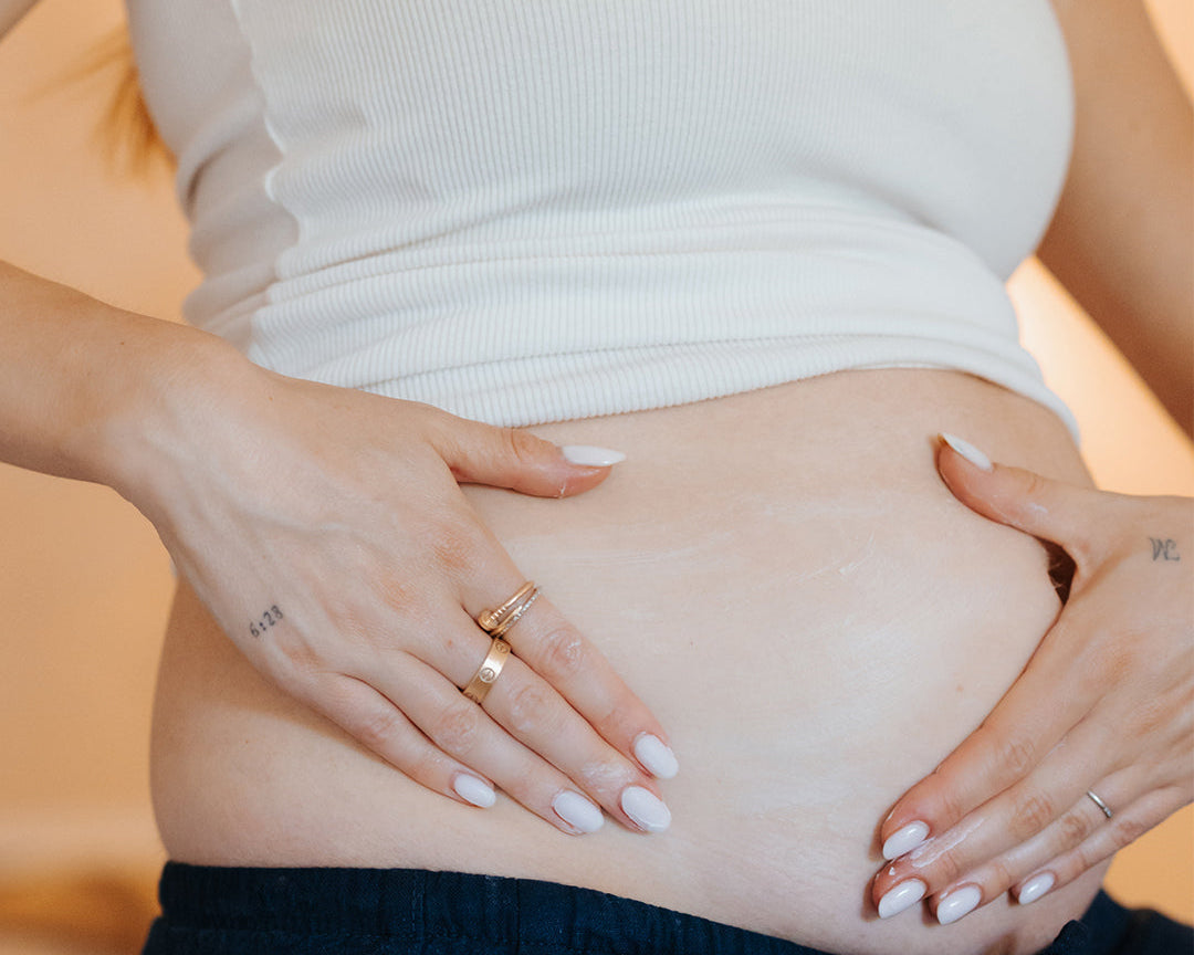 A close-up lifestyle shot of a pregnant woman wearing a white ribbed tank top and dark navy pants, cradling her baby bump with her hands. She is wearing gold rings, and the background is a soft, warm beige.