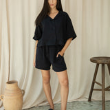 Woman in a black outfit standing against a beige curtain with a wooden stool and vase in the background.