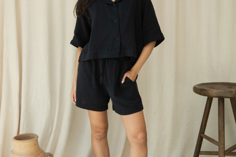 Woman in a black outfit standing against a beige curtain with a wooden stool and vase in the background.