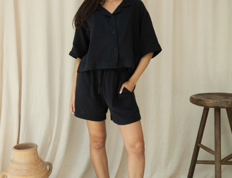 Woman in a black outfit standing against a beige curtain with a wooden stool and vase in the background.
