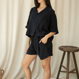 Woman wearing a black outfit standing against a beige curtain with a wooden stool.