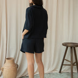 Woman wearing a black outfit standing against a beige curtain with a vase and stool in the foreground.