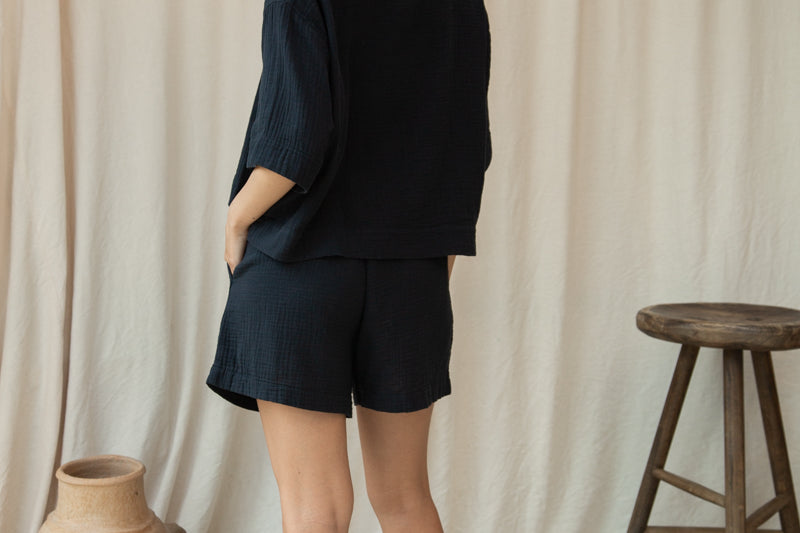 Woman wearing a black outfit standing against a beige curtain with a vase and stool in the foreground.
