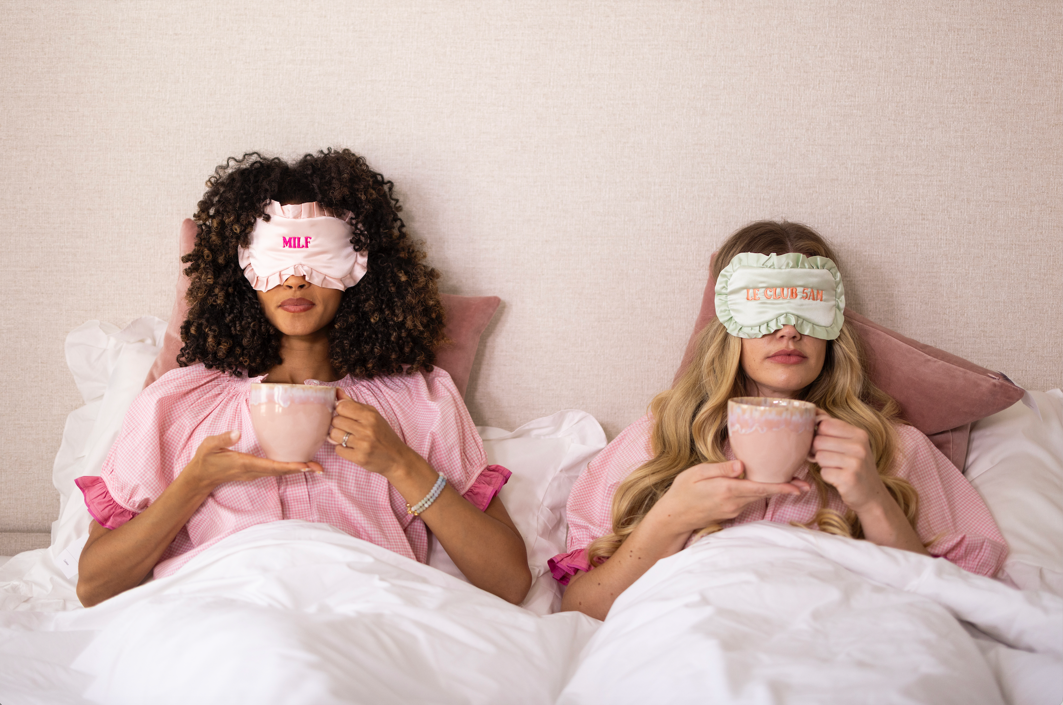 Two women in pink robes lying in bed under white covers, each wearing a sleep mask. The mask on the left is light pink with the text 'MILF', and the mask on the right is light green with the text 'LE CHAMBER'.