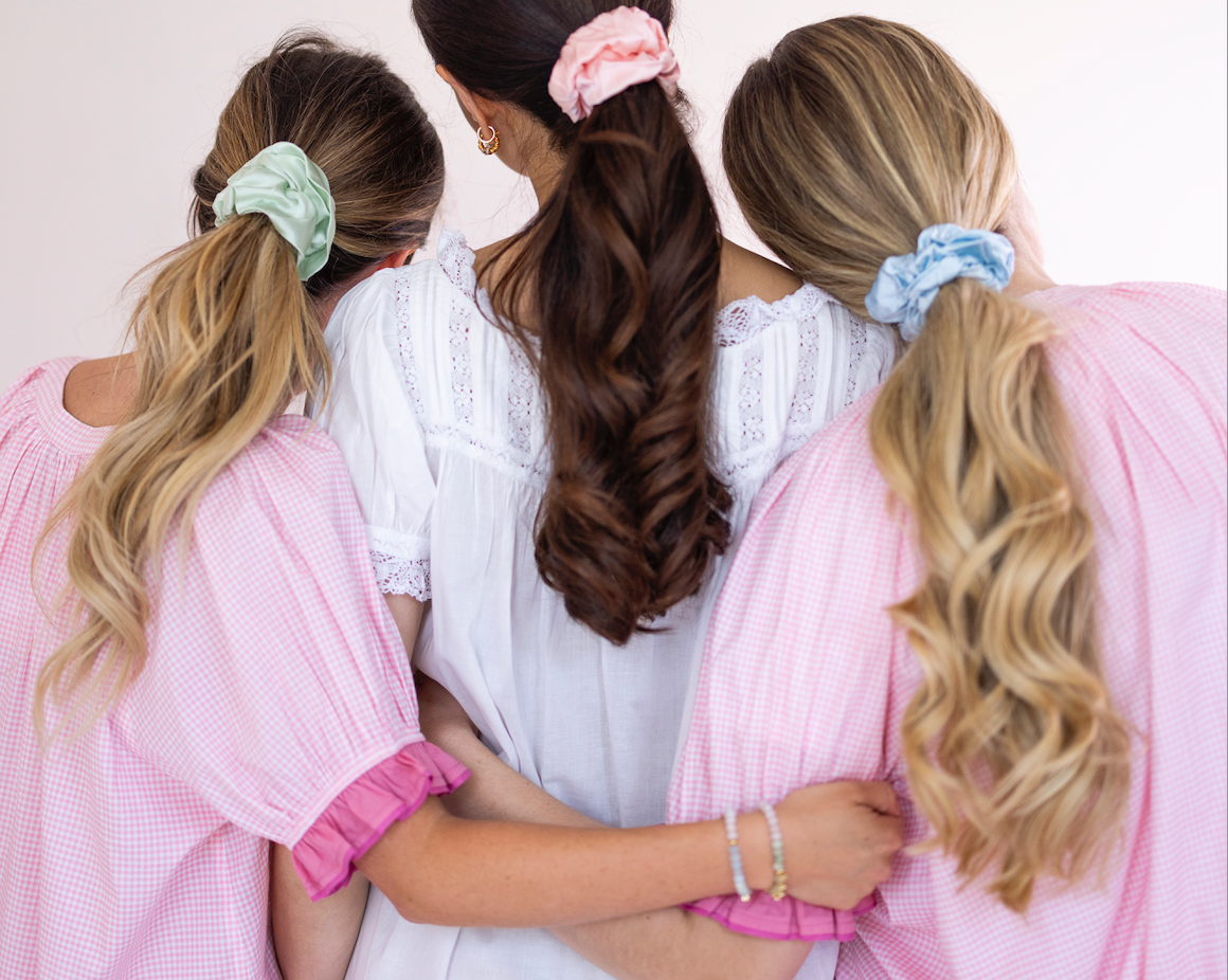 Three women standing together from behind, wearing matching pink gingham dresses and hair accessories. The center woman wears a white dress, while the two on the sides wear pink dresses.